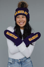A smiling woman stands against a gray background with her arms crossed, wearing a purple beanie and the Panache Apparel Collection’s Purple & Gold Game Day Mittens—cozy, fleece-lined knit mittens featuring "GAMEDAY" lettering.