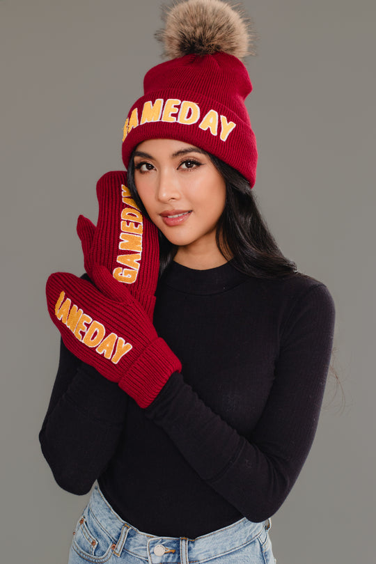 A woman in a black turtleneck and jeans wears a maroon & gold Game Day Pom Hat from the Panache Apparel Collection, along with matching mittens, both featuring "GAMEDAY" in yellow letters. She holds one mitten to her cheek and smiles softly.