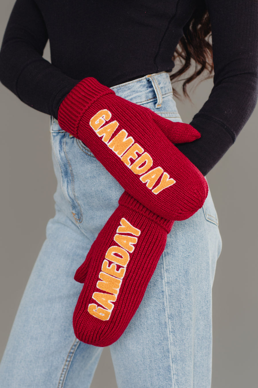 A person wearing blue jeans and a black top stands with their arms crossed, displaying the Maroon & Gold Game Day Mittens from Panache Apparel Collection. These fleece-lined maroon knit mittens feature "GAMEDAY" in bold yellow letters for extra warmth and style, set against a plain gray background.