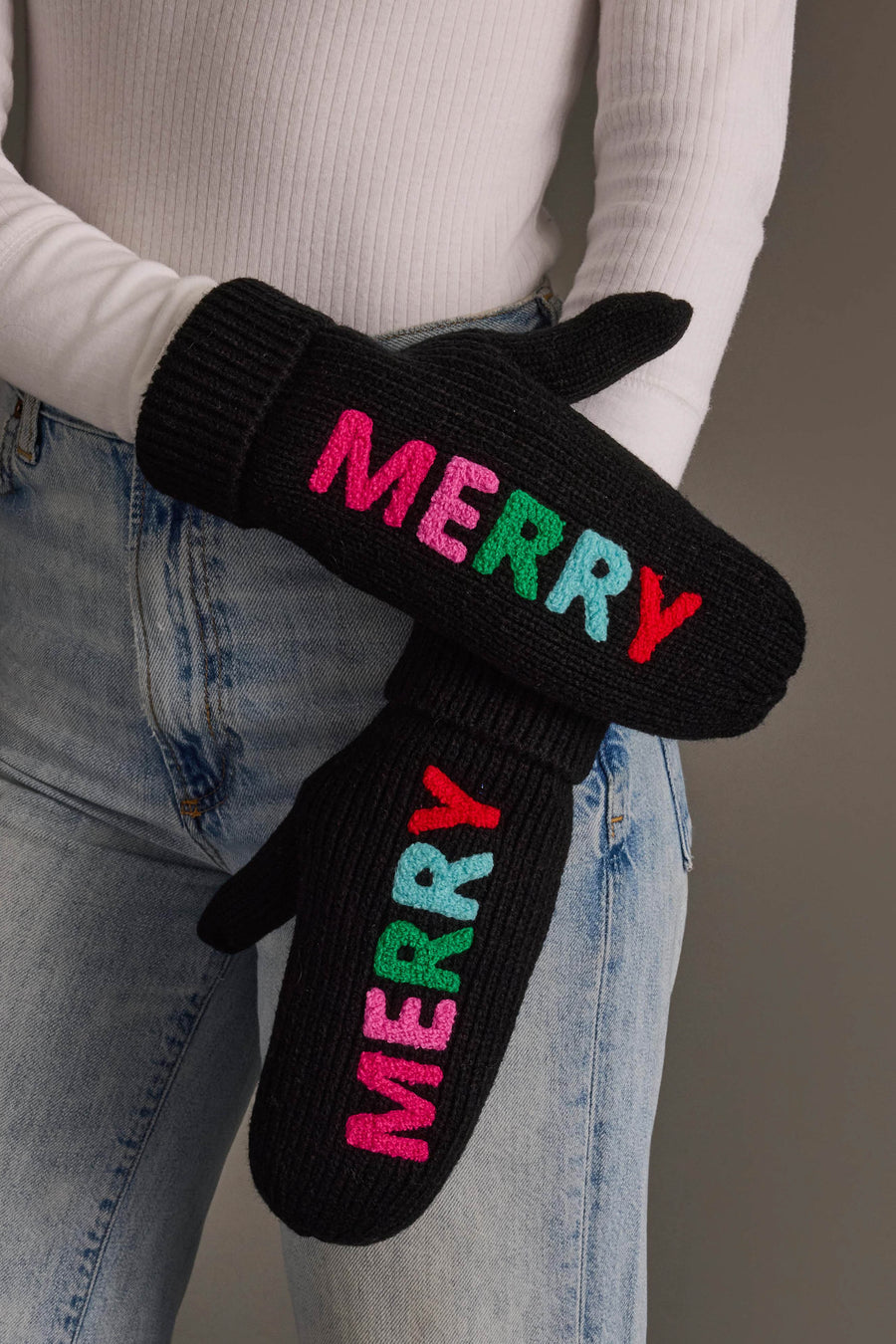 A person wearing Black Merry Knit Mittens, featuring the word MERRY embroidered in colorful lettering on each black acrylic knit mitten.