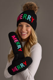 A person wears a black knit beanie with a faux fur pompom and matching Black Merry Knit Mittens from the Panache Apparel Collection, all featuring the word MERRY in multi-colored letters.