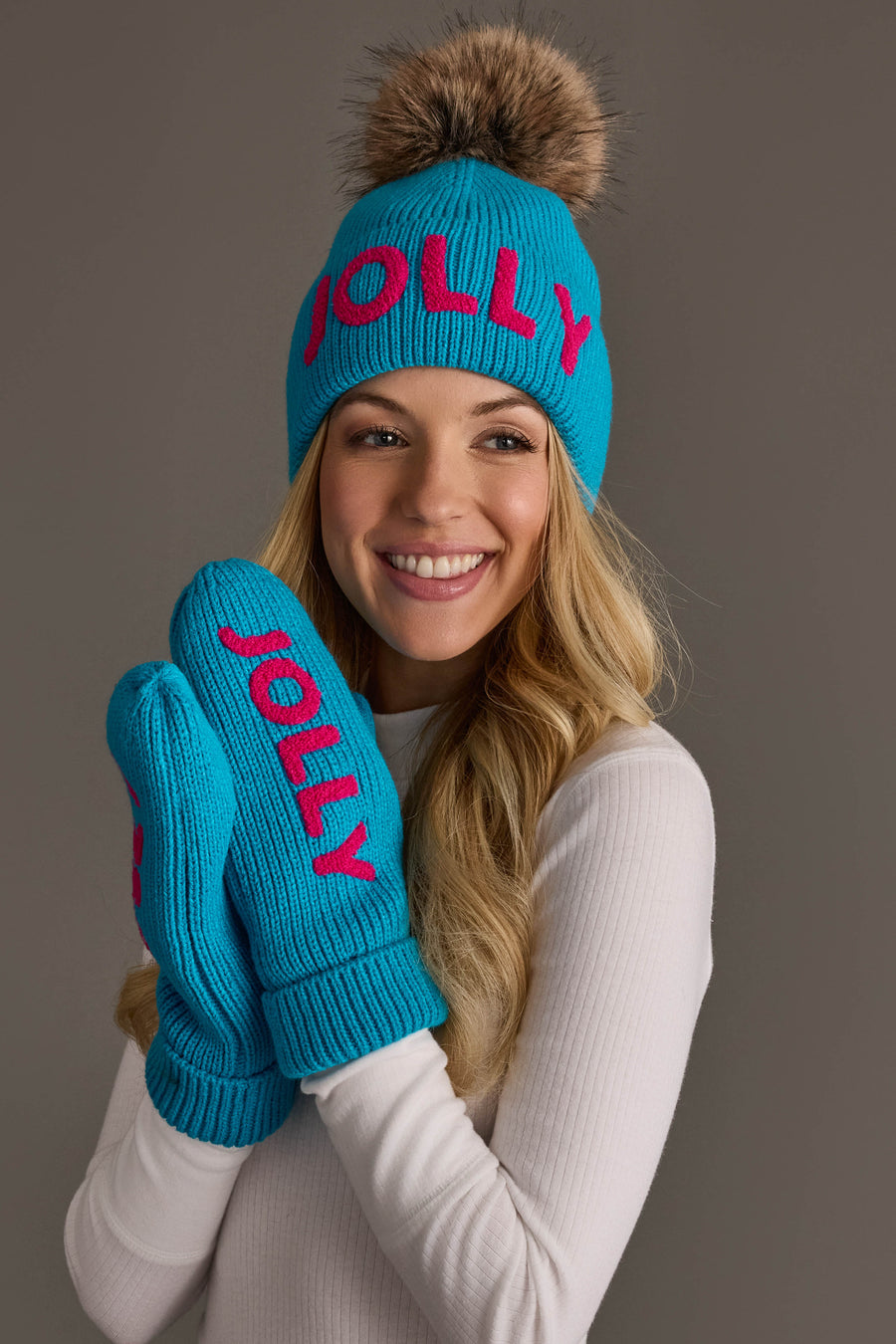 A smiling woman wears a Blue Jolly Knit Pom Hat with a brown faux-fur pompom and matching blue mittens, both featuring the word JOLLY in pink text.