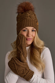 A woman with long blonde hair smiles as she poses in a Panache Apparel Collection brown cable knit hat with a dark orange pom and matching mittens, holding one mitten to her face. She is dressed in a white ribbed turtleneck against a plain background.