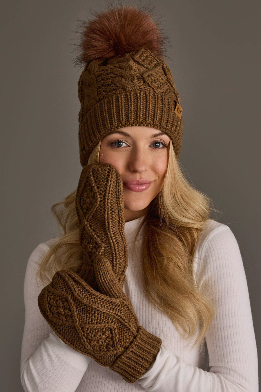 A woman with long blonde hair smiles as she poses in a Panache Apparel Collection brown cable knit hat with a dark orange pom and matching mittens, holding one mitten to her face. She is dressed in a white ribbed turtleneck against a plain background.