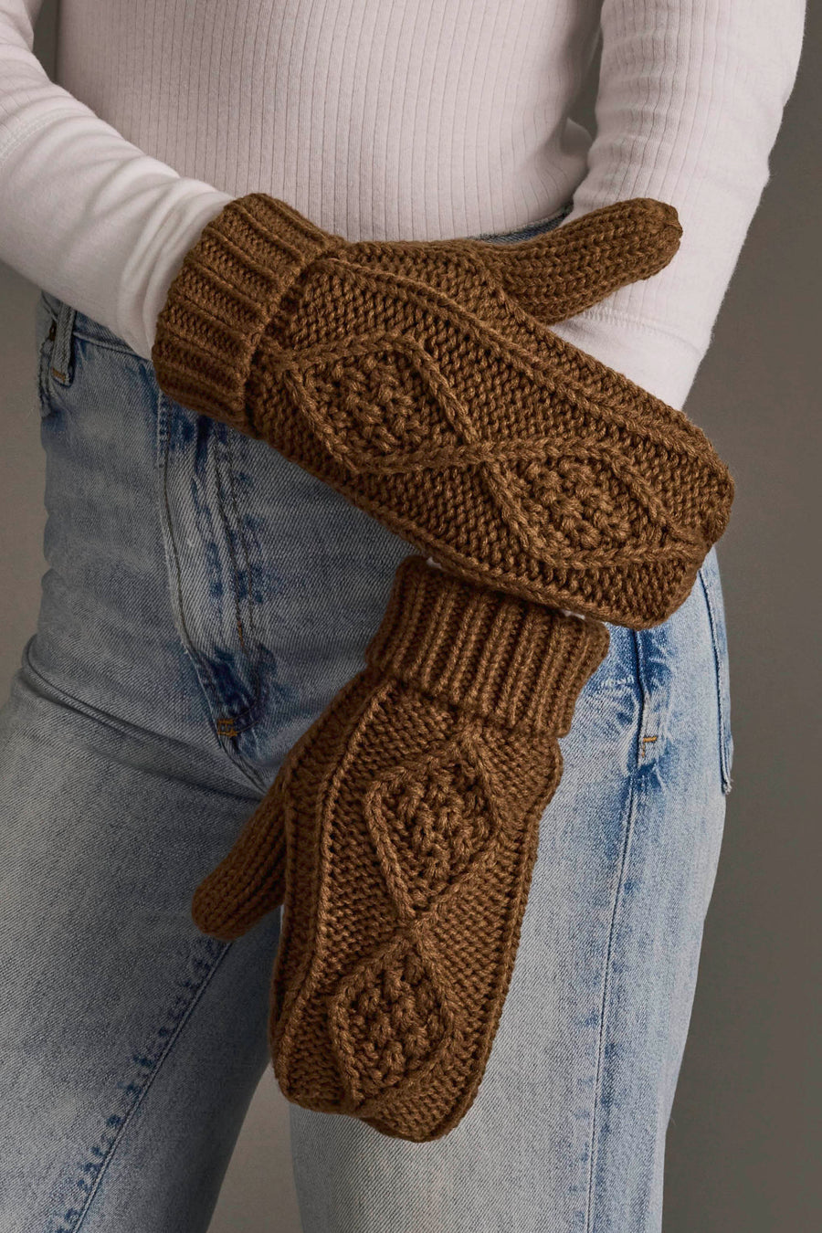 A person wearing a white ribbed long-sleeve top, light blue jeans, and the Brown Cable Knit Mittens from Panache Apparel Collection, featuring a chunky knit and diamond pattern, stands with their hands crossed at the waist.