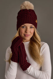A woman in a white turtleneck wears the Panache Apparel Collection’s Burgundy Cable Knit Hat with Light Tan Pom and matching mittens, smiling softly in front of a plain gray background.