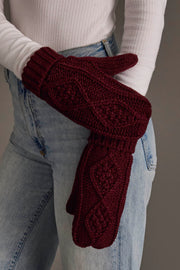 A person wearing a white ribbed sweater and blue jeans has their arms crossed, showing off the Panache Apparel Collection’s Burgundy Cable Knit Mittens, featuring a chunky texture and intricate patterns.