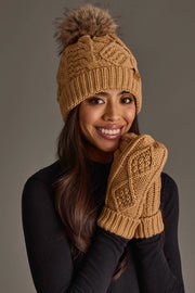 A smiling woman wears the Panache Apparel Collection Khaki Cable Knit Pom Hat with matching brown knit mittens and a black long-sleeve top, posing against a plain background.