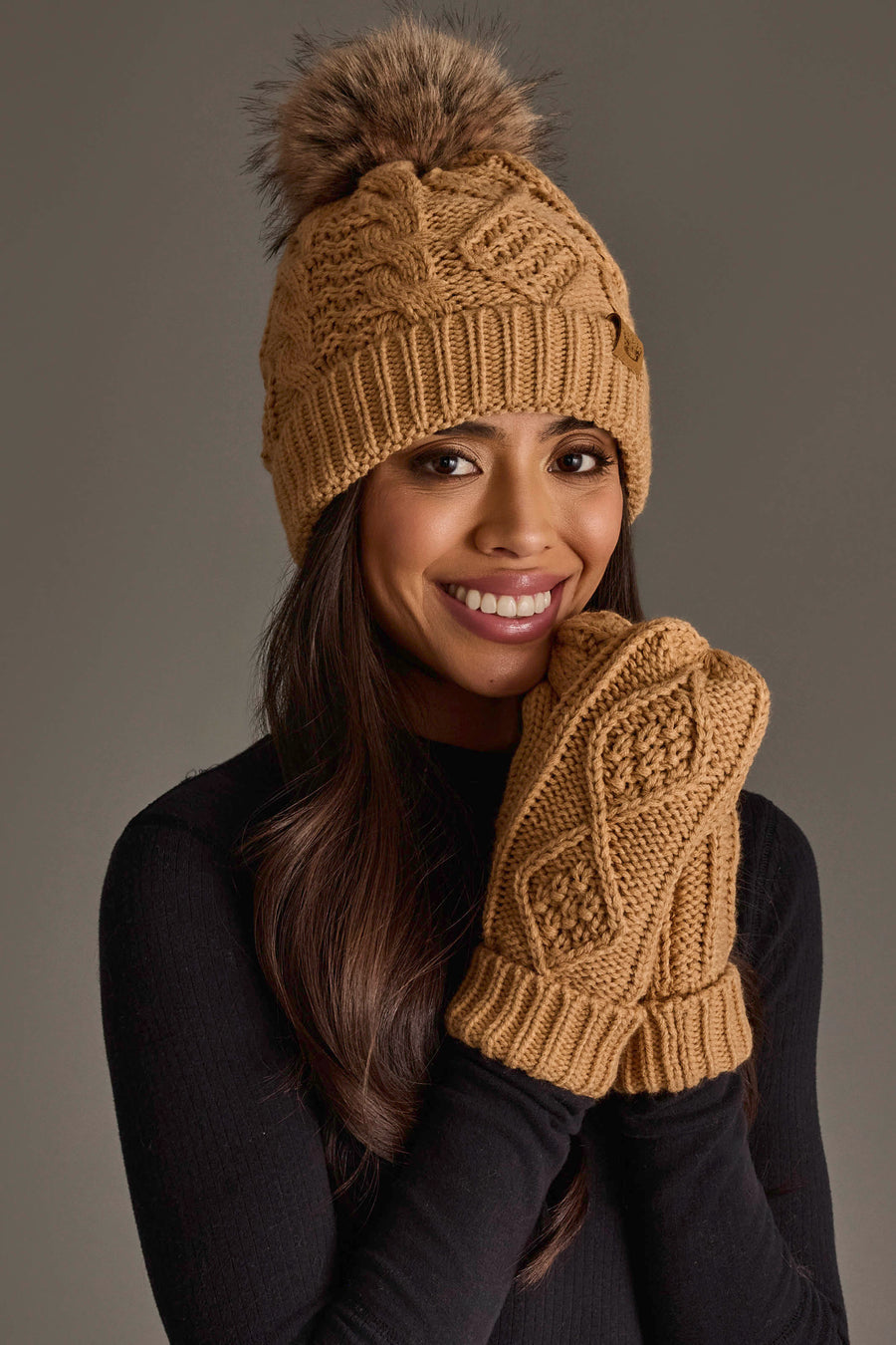 A smiling woman wears the Panache Apparel Collection Khaki Cable Knit Pom Hat with matching brown knit mittens and a black long-sleeve top, posing against a plain background.