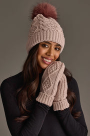 A woman with long brown hair and a black top smiles against a plain background, holding her hands, dressed in matching mittens, to her face as she wears the Light Pink Cable Knit Hat with Dark Pink Pom by Panache Apparel Collection.