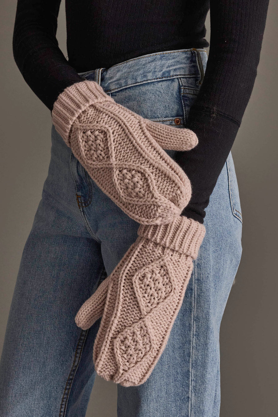 A person in blue jeans and a black top wears light pink cable knit mittens from Panache Apparel Collection.