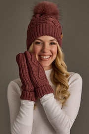 A smiling woman in a white sweater wears the Panache Apparel Collection Dark Pink Cable Knit Hat with Dark Pink Pom and matching mittens, holding her hands up to her face against a neutral gray background.