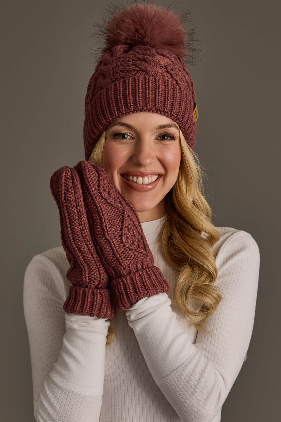 A smiling woman in a white sweater wears the Panache Apparel Collection Dark Pink Cable Knit Hat with Dark Pink Pom and matching mittens, holding her hands up to her face against a neutral gray background.