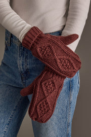 A person wearing a white ribbed turtleneck and blue jeans holds a pair of Panache Apparel Collection Dark Pink Cable Knit Mittens, featuring a raised diamond pattern across the back.