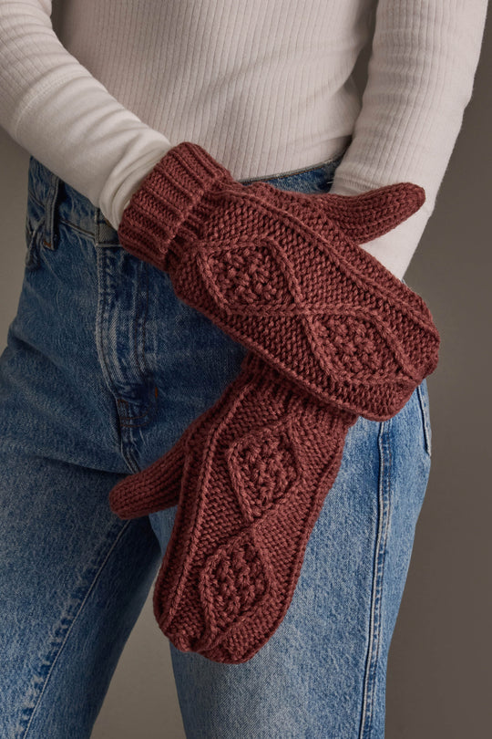 A person wearing a white ribbed turtleneck and blue jeans holds a pair of Panache Apparel Collection Dark Pink Cable Knit Mittens, featuring a raised diamond pattern across the back.