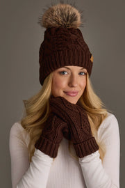 A woman with long blonde hair wears the Panache Apparel Collection’s Dark Brown Cable Knit Pom Hat, paired with matching brown knit gloves. She smiles softly in a white long-sleeve top against a plain gray background.