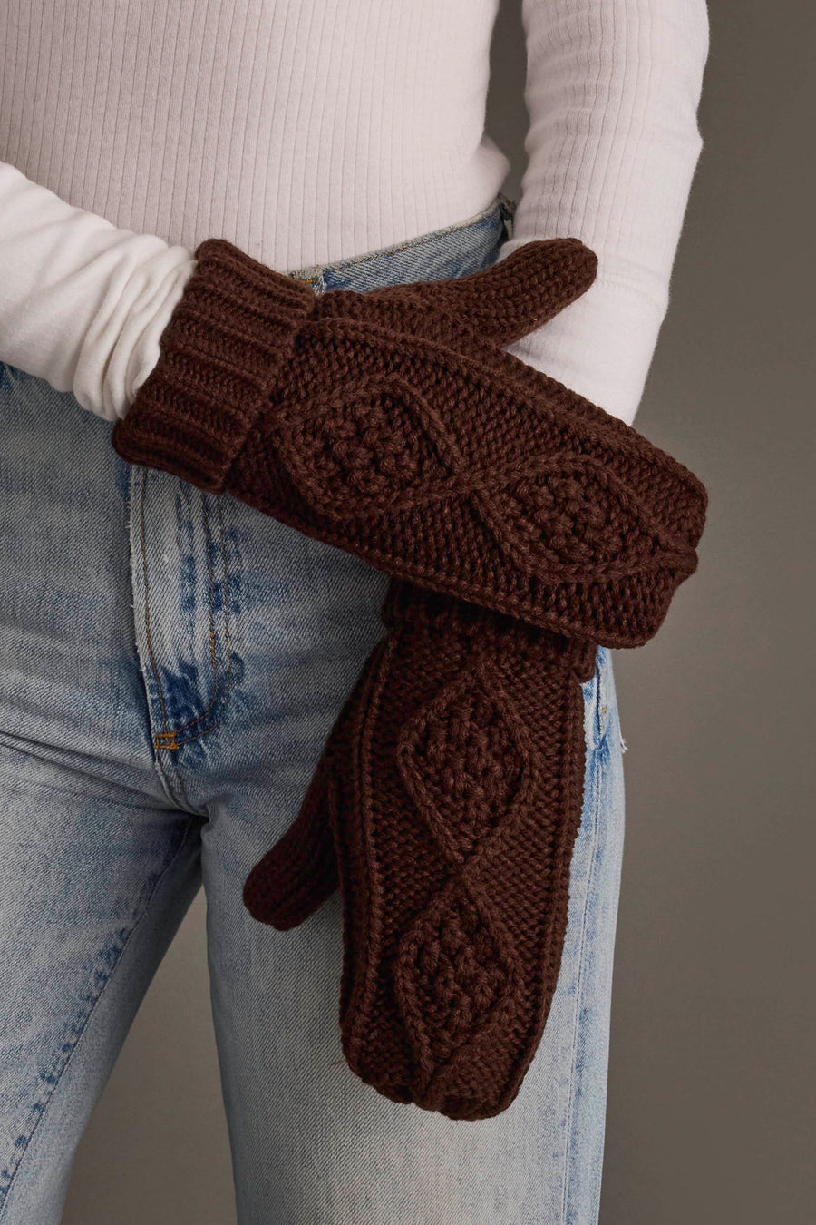 A person wearing light wash jeans and a white long-sleeve shirt holds a pair of Panache Apparel Collection Dark Brown Cable Knit Mittens.