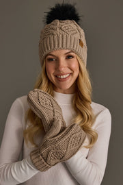 A woman wearing the Panache Apparel Collection Warm Taupe Cable Knit Pom Hat smiles at the camera. The hat features a black pom-pom and is paired with matching beige mittens. She has long blonde hair and is dressed in a white long-sleeve top.