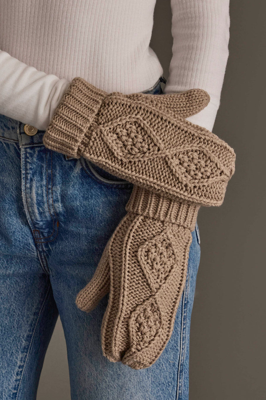 A person wearing blue jeans and a white sweater holds the Warm Taupe Cable Knit Mittens by Panache Apparel Collection.