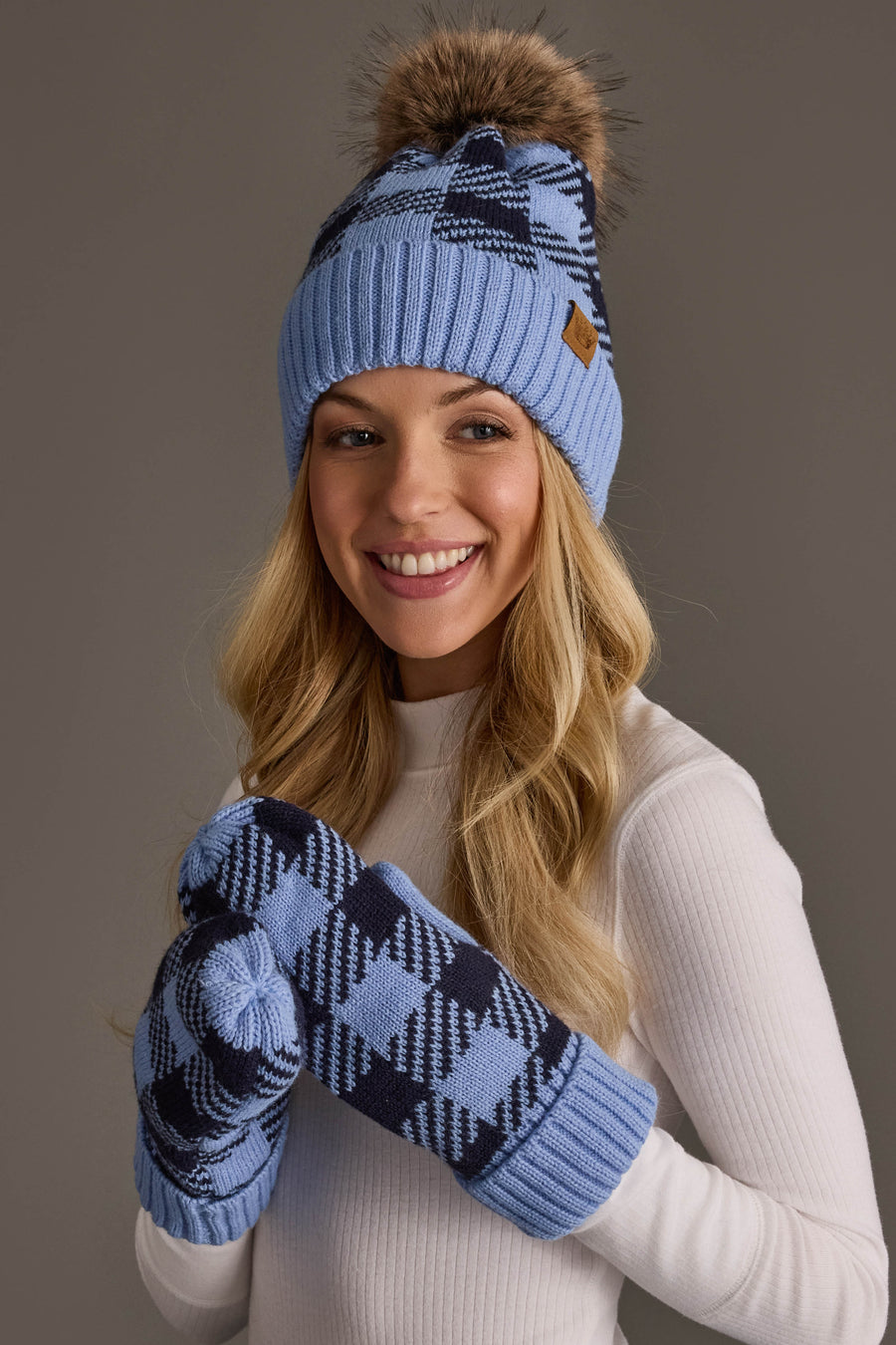 A smiling woman in a white top wears the Panache Apparel Collection Light Blue & Navy Plaid Knit Pom Hat, paired with matching mittens, standing against a plain gray background.