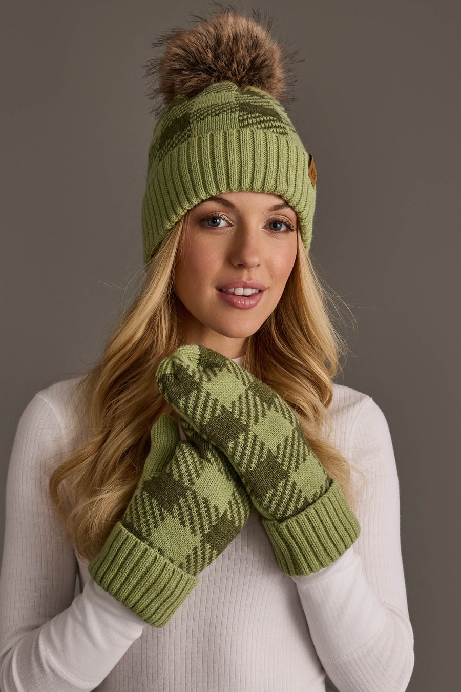 A woman with long blonde hair wears the Panache Apparel Collection Light Green & Olive Plaid Knit Pom Hat, featuring a faux fur pom-pom, and poses against a neutral background in a white ribbed top and coordinating green patterned mittens.