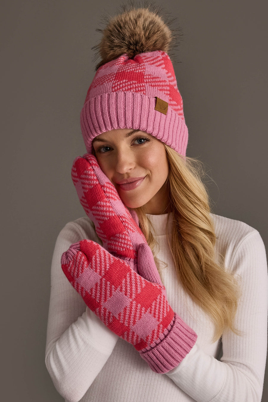 A woman wears the Panache Apparel Collection Pink & Bright Pink Plaid Knit Pom Hat, smiling gently while holding her hands, covered in matching pink patterned mittens, near her face against a plain background.