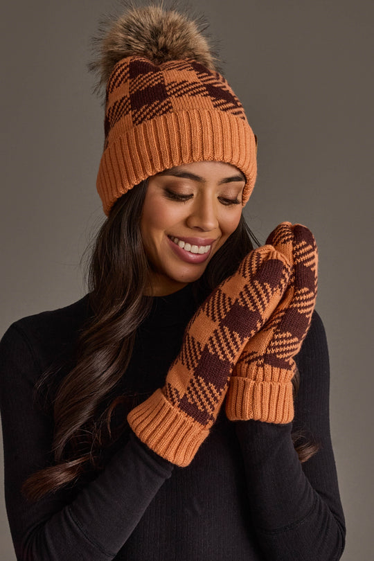 A woman wearing the Panache Apparel Collection Orange & Brown Plaid Knit Pom Hat, smiling with her eyes closed, pairs it with matching mittens and a black top.