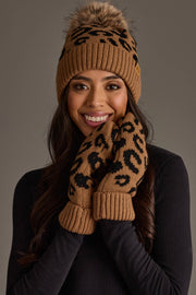 A woman smiles while wearing the Panache Apparel Collection Camel Leopard Pattern Knit Pom Hat and coordinating brown and black leopard print mittens, posing against a gray background.