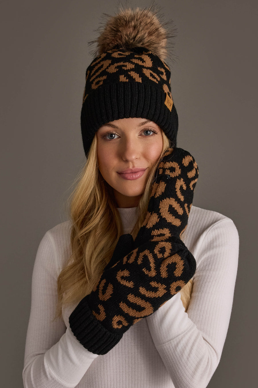 A woman in a white sweater wears the Panache Apparel Collection’s Black Leopard Pattern Knit Pom Hat, featuring a fur pom-pom, along with matching black and brown leopard print mittens, smiling softly against a plain background.
