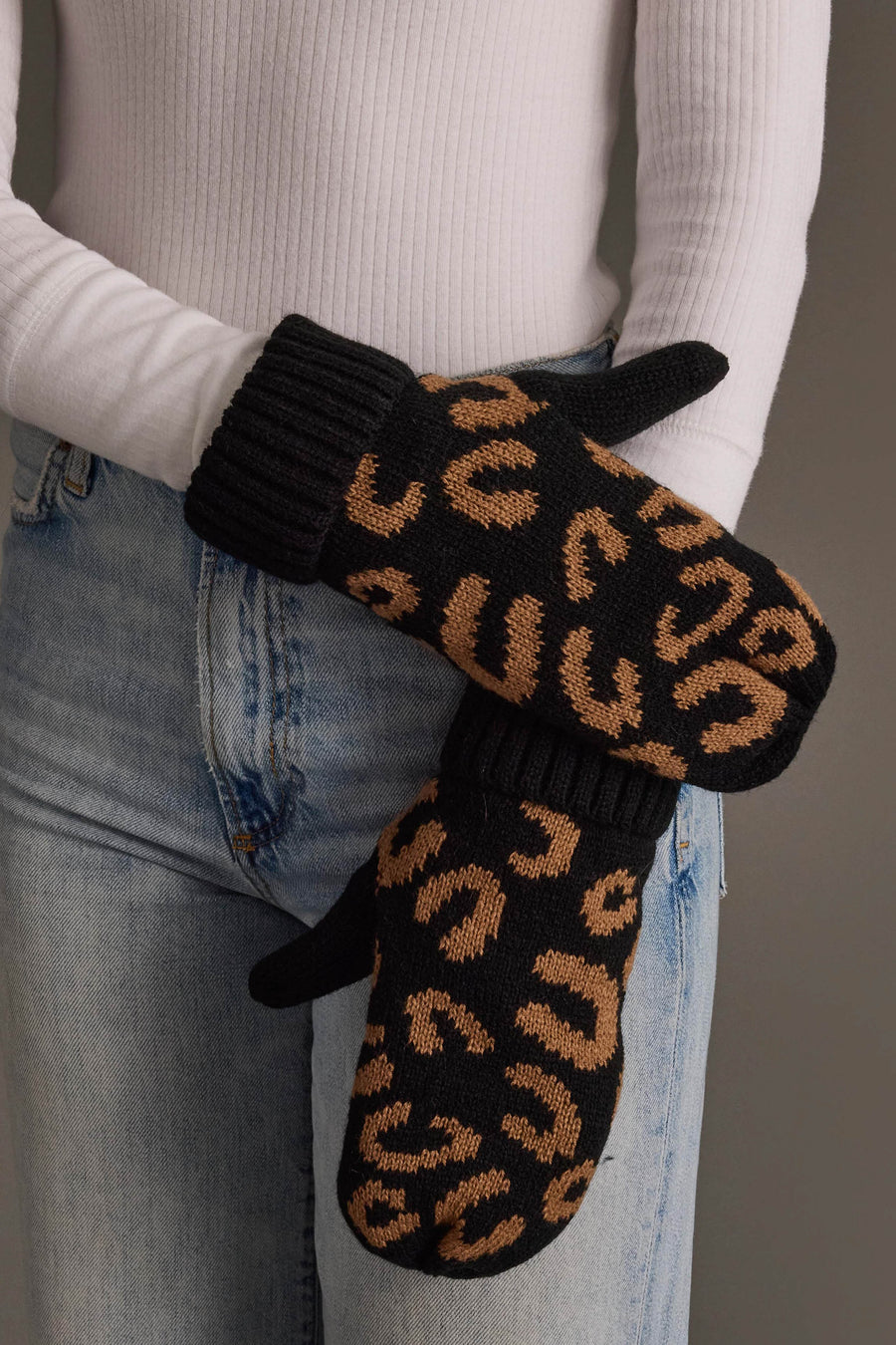 A person wearing blue jeans, a white ribbed sweater, and Panache Apparel Collection Black Leopard Pattern Knit Mittens crosses their arms in front of their body.