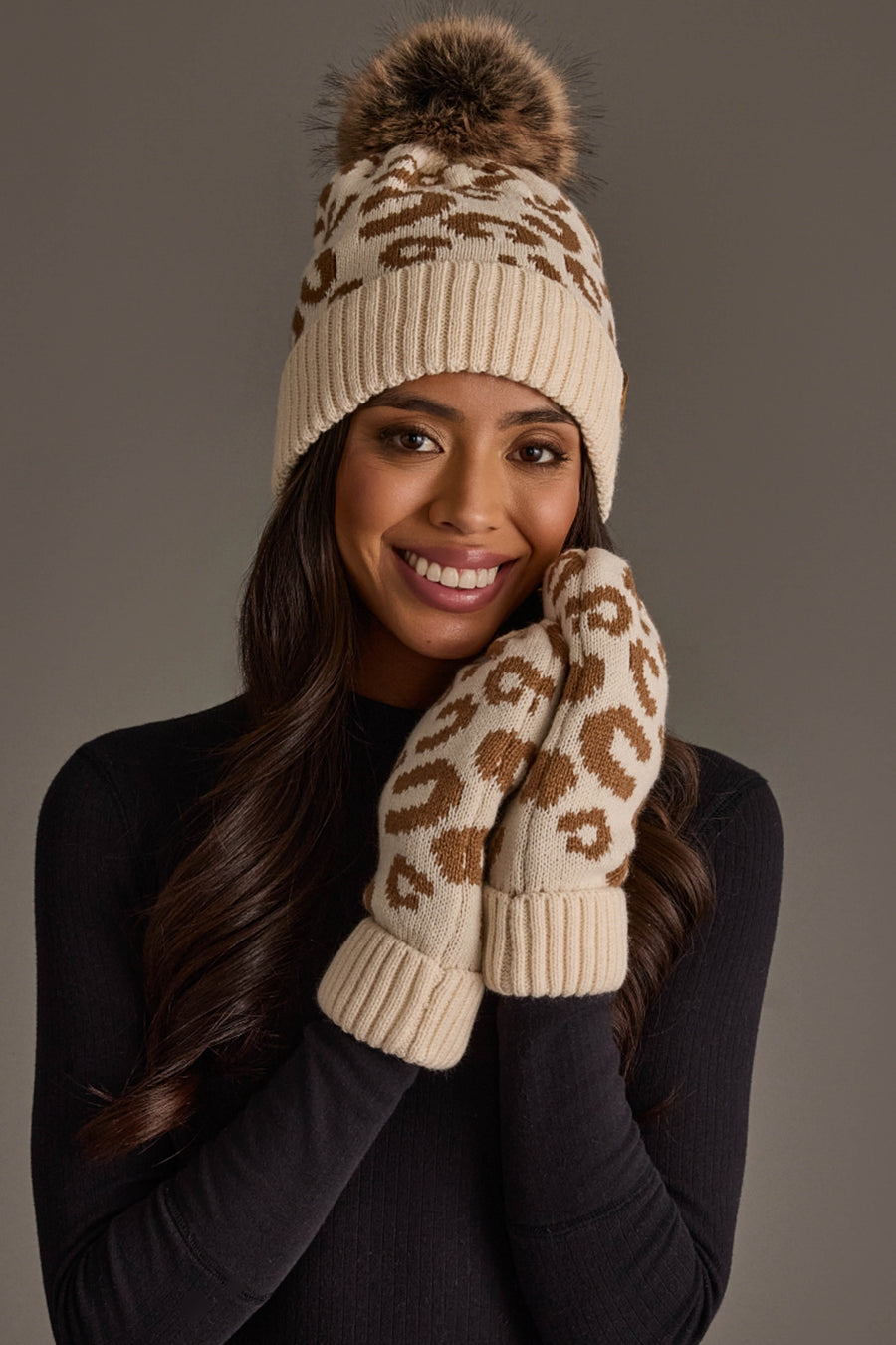 A woman in a black top smiles against a neutral background, wearing the Panache Apparel Collection’s Cream Leopard Pattern Knit Pom Hat and matching mittens, both featuring a brown leopard print design.