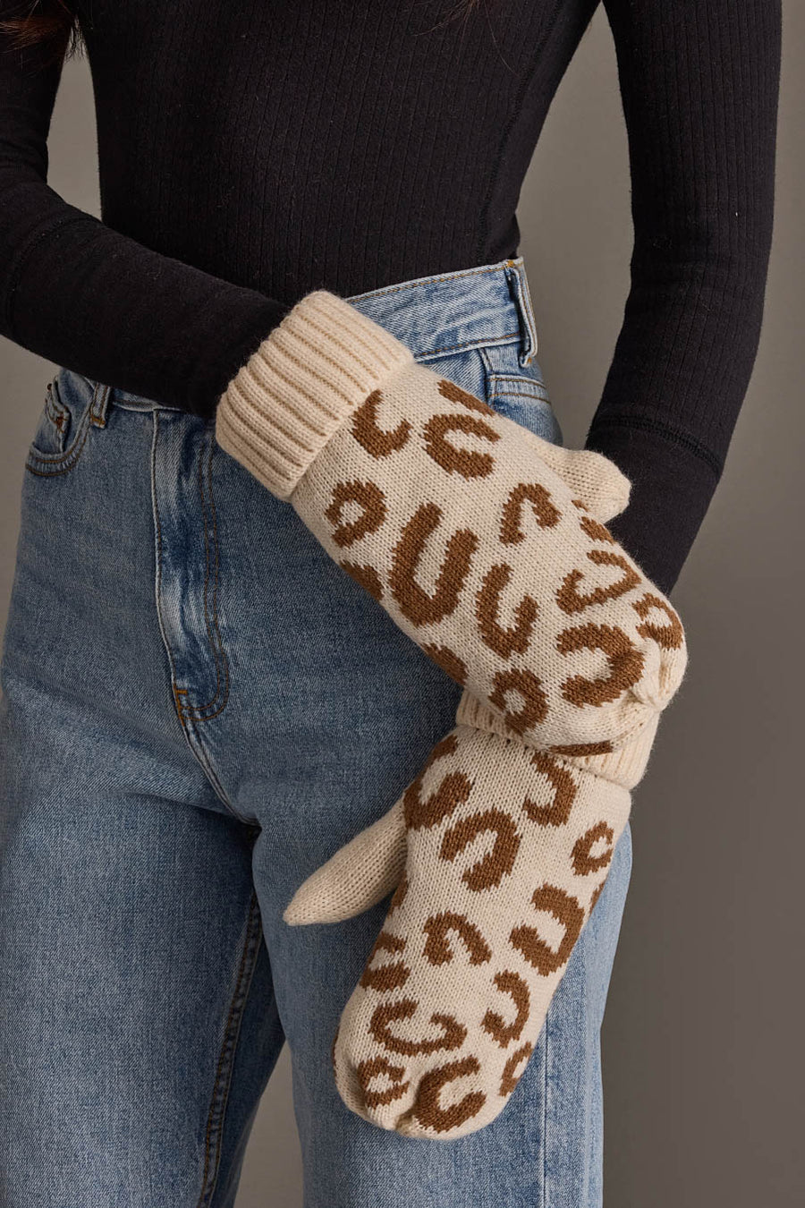 A person in blue jeans and a black ribbed top holds the Cream Leopard Pattern Knit Mittens from Panache Apparel Collection.