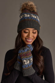 A woman smiles as she looks down, wearing the Panache Apparel Collection Gray Swan Pattern Knit Pom Hat, with a fur pom-pom, and matching mittens featuring a colorful animal-themed design, set against a plain gray background.