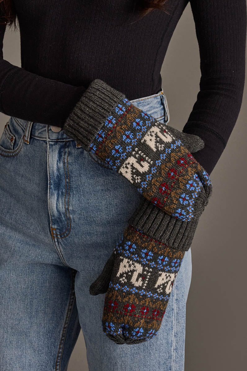 A person wearing blue jeans, a black ribbed long-sleeve shirt, and Panache Apparel Collection’s Gray Swan Pattern Knit Mittens, featuring a colorful Nordic design in gray, white, blue, red, and green, stands against a neutral background.