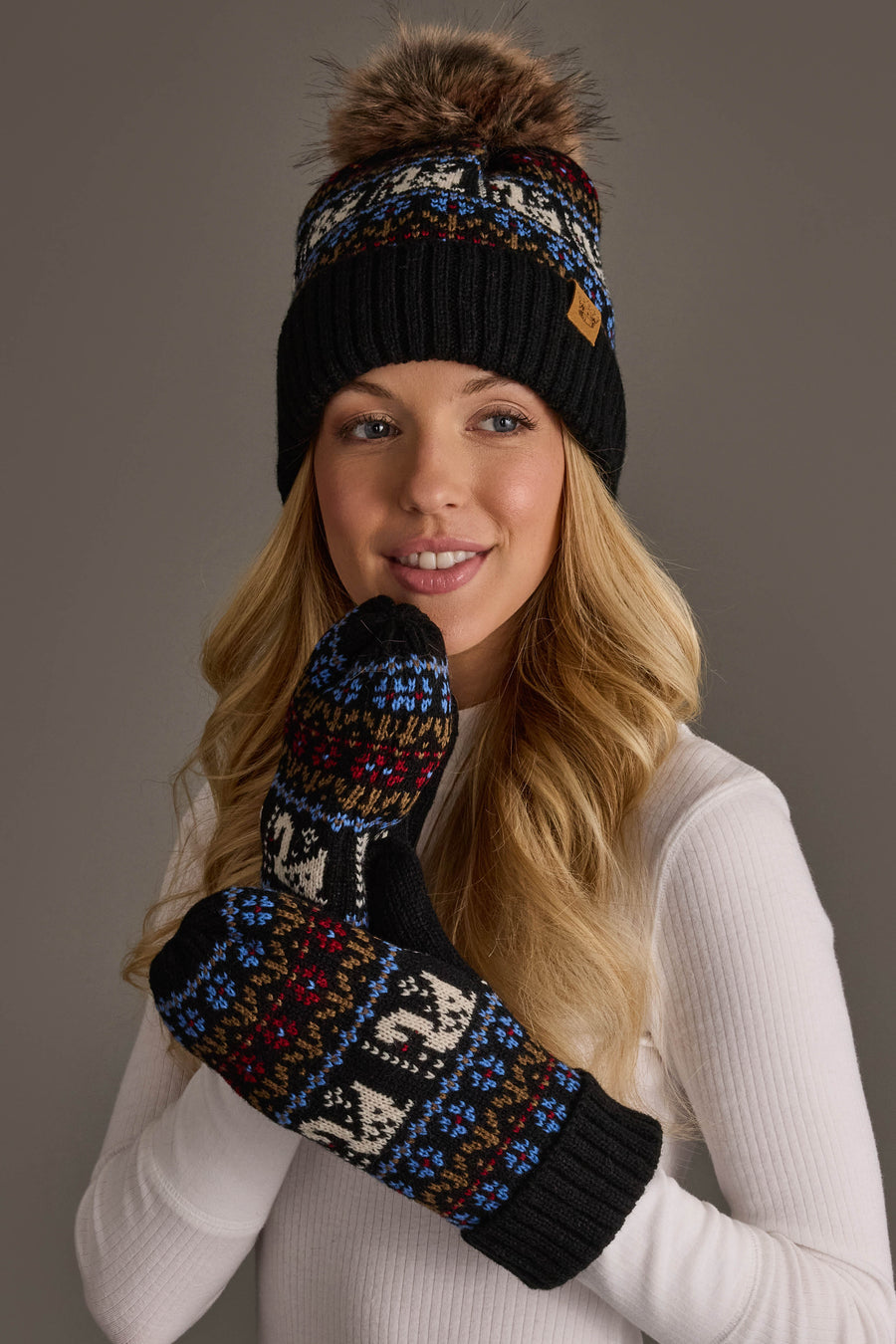 A woman in a white turtleneck sweater smiles against a plain gray background while wearing the Panache Apparel Collection’s Black Swan Pattern Knit Pom Hat and matching patterned mittens.
