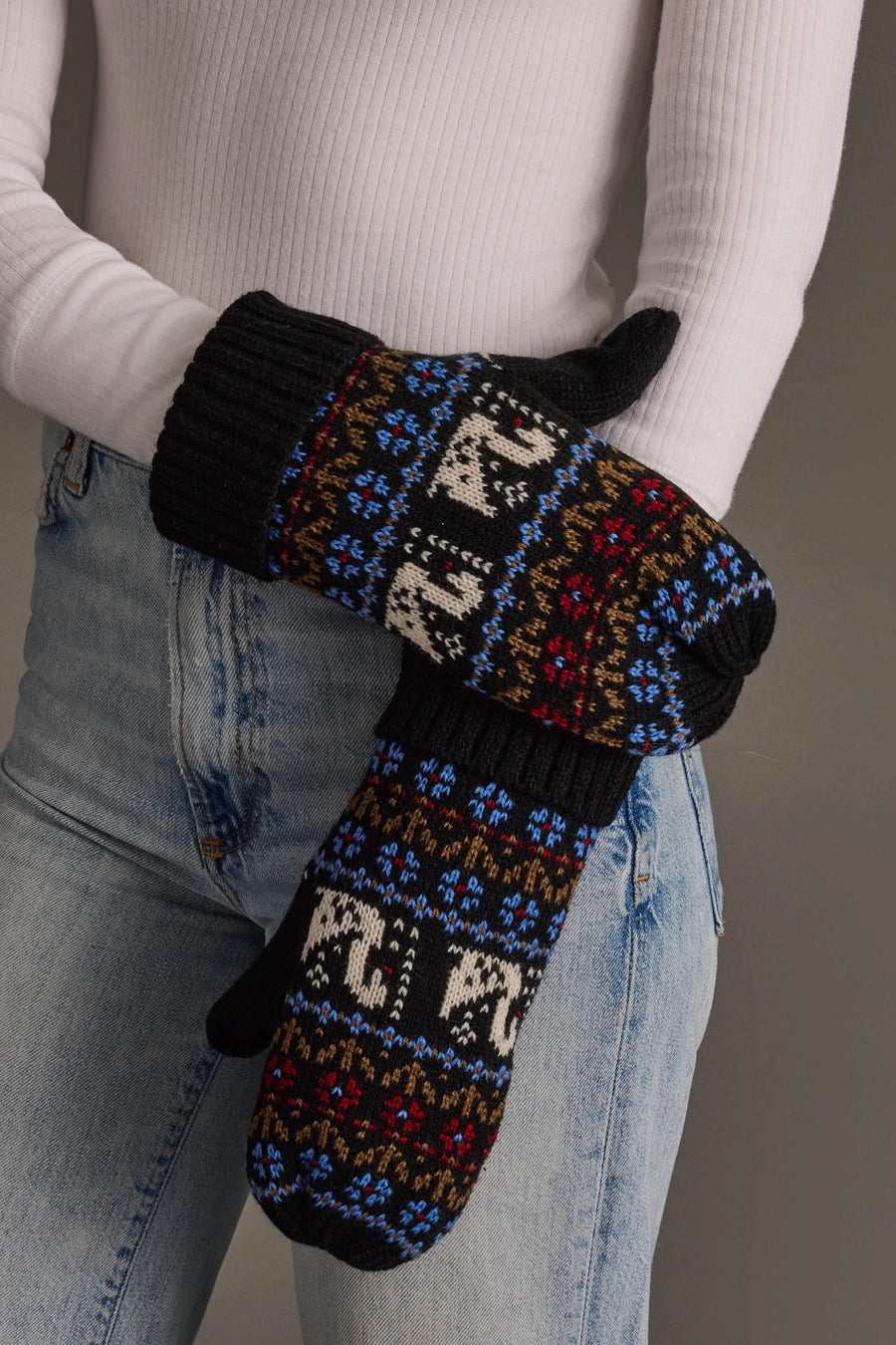 A person wearing light blue jeans, a white ribbed sweater, and Panache Apparel Collection’s Black Swan Pattern Knit Mittens, featuring colorful black, blue, brown, red, and white designs, crosses their arms.