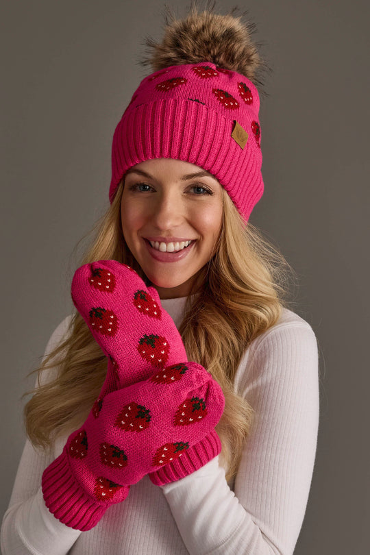 A smiling person in a white top poses against a plain gray background, wearing the Panache Apparel Collection’s Strawberry Pattern Pom Hat, a bright pink knit hat with a pom-pom, along with matching mittens, both adorned with charming strawberry designs.