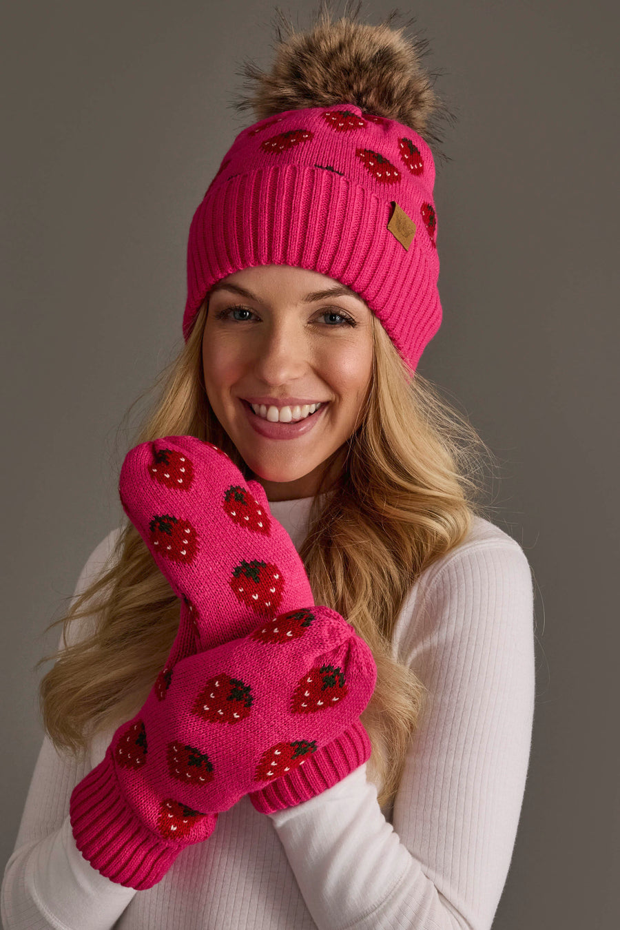 A smiling person in a white top poses against a plain gray background, wearing the Panache Apparel Collection’s Strawberry Pattern Pom Hat, a bright pink knit hat with a pom-pom, along with matching mittens, both adorned with charming strawberry designs.