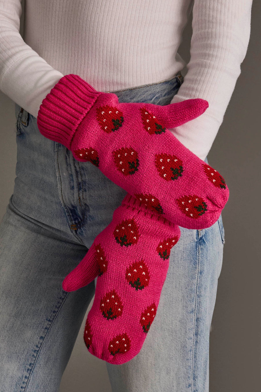 A person wearing the Panache Apparel Collection Strawberry Pattern Mittens, featuring a bright pink color with red strawberry designs, paired with a white ribbed sweater and blue jeans. Their arms are crossed against a plain, neutral background.