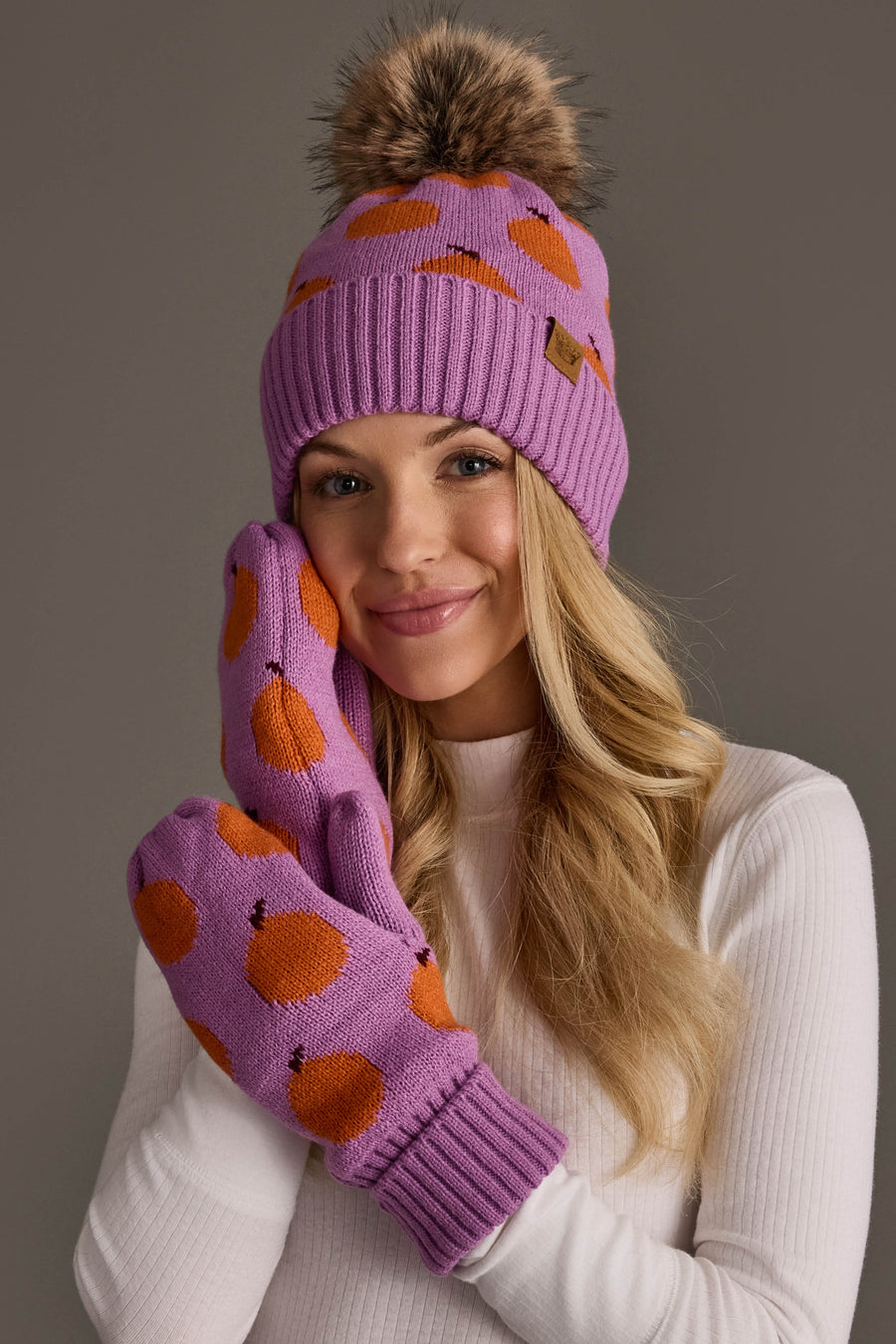 A woman smiles and poses with her hand to her face, wearing the Panache Apparel Collection Orange Pattern Pom Hat, a purple beanie with orange spots and a pom-pom, paired with matching mittens and a white ribbed shirt against a plain background.