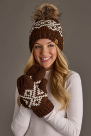 A smiling woman in a white sweater wears the Panache Apparel Collection’s Brown & White Patterned Pom Hat, paired with matching mittens, against a plain gray background.