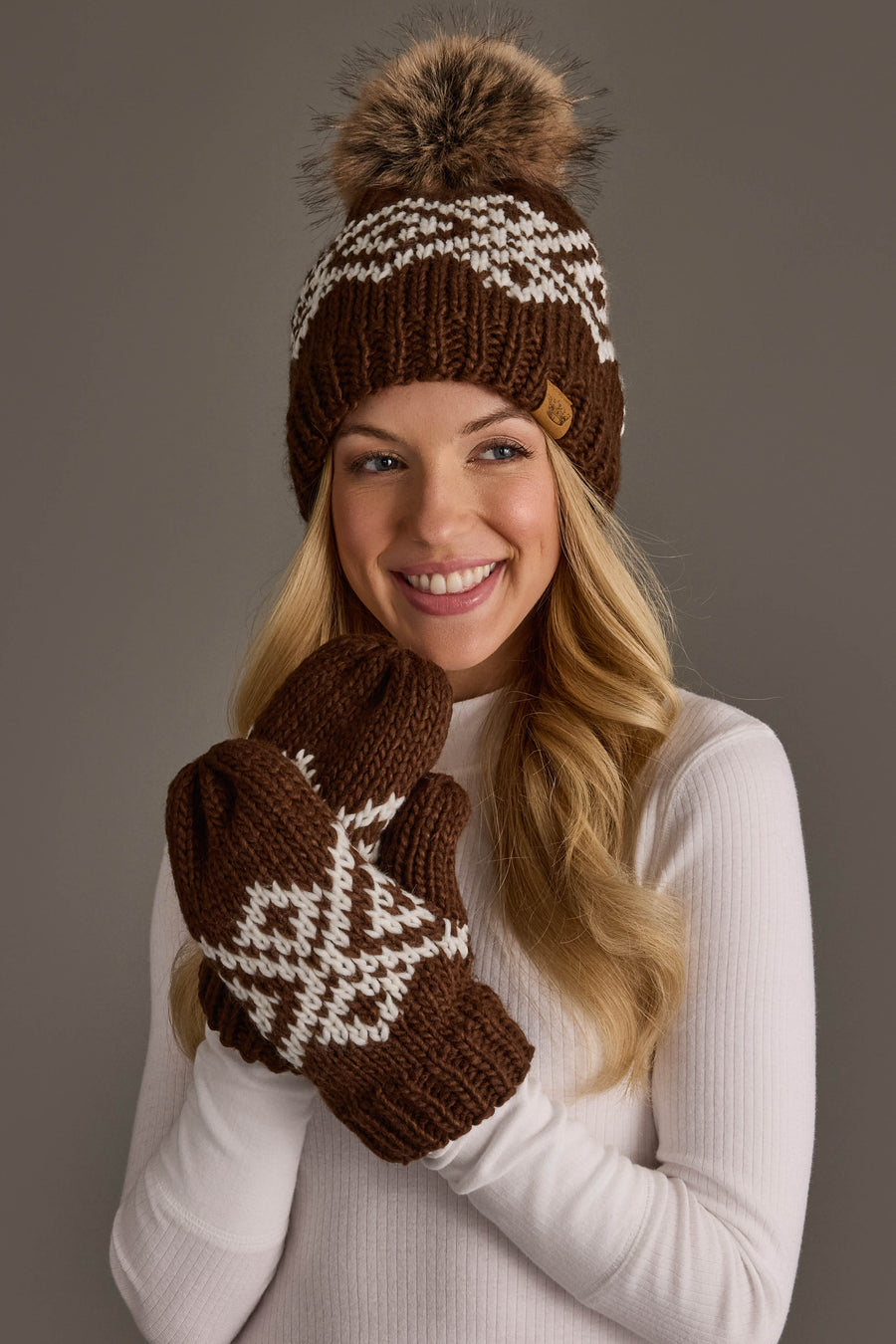 A smiling woman in a white sweater wears the Panache Apparel Collection’s Brown & White Patterned Pom Hat, paired with matching mittens, against a plain gray background.