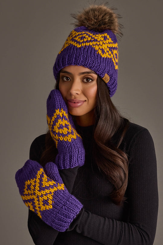 A woman models the Panache Apparel Collection’s Purple & Gold Patterned Pom Hat and matching mittens, wearing her long hair down with a black sweater. She poses with one hand to her face against a neutral background.