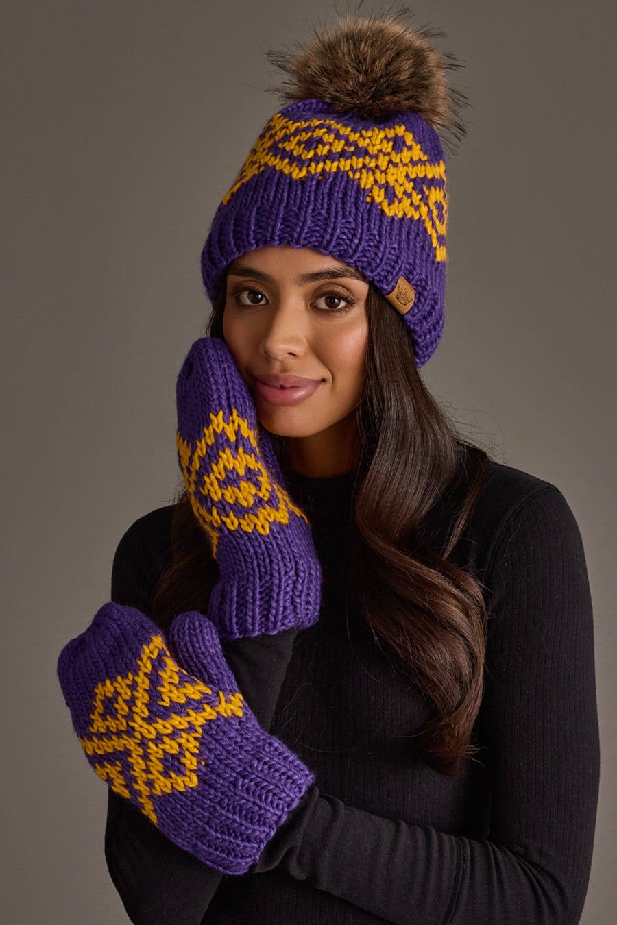 A woman models the Panache Apparel Collection’s Purple & Gold Patterned Pom Hat and matching mittens, wearing her long hair down with a black sweater. She poses with one hand to her face against a neutral background.
