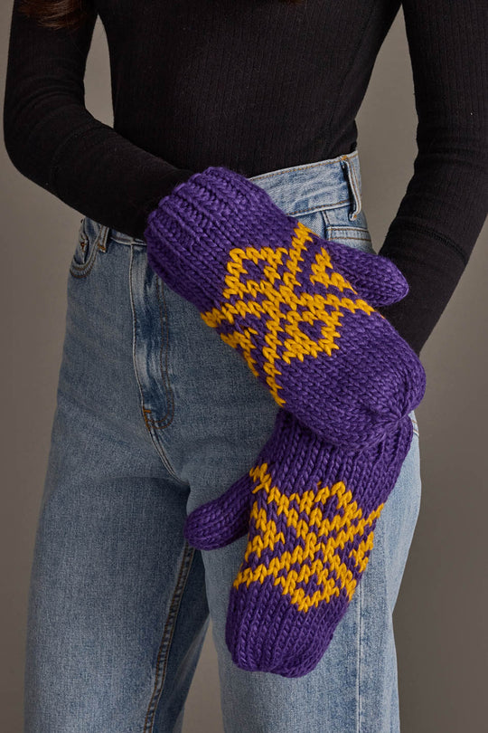 A person wearing a black long-sleeve top and blue jeans holds the Purple & Gold Patterned Mittens from Panache Apparel Collection.