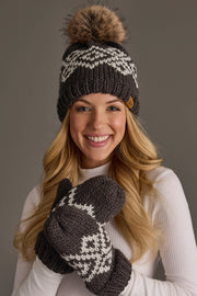 A woman smiles while posing against a plain gray background, wearing the Panache Apparel Collection Dark Gray & White Patterned Pom Hat, styled with matching mittens and a white ribbed turtleneck sweater.