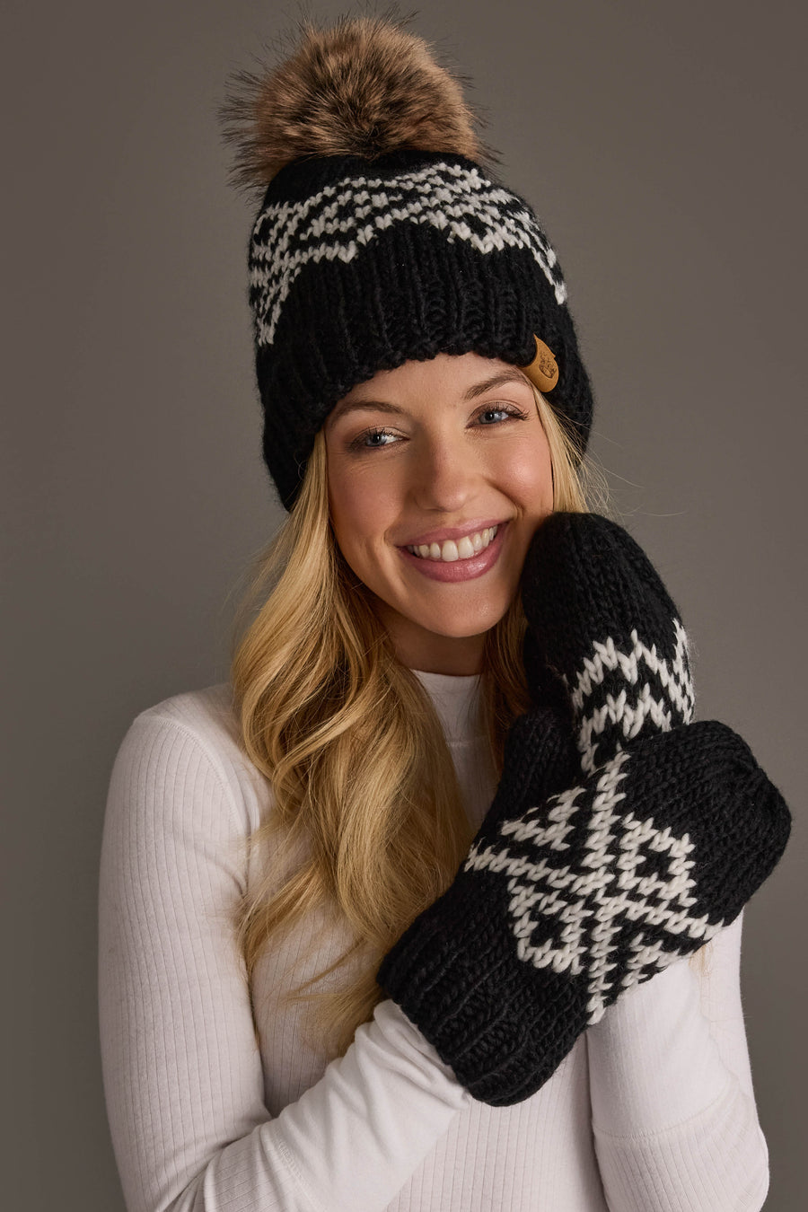 A smiling woman stands against a gray background, wearing matching Panache Apparel Collection Black & White Patterned Mittens, a white sweater, and a black knit hat with a pompom.