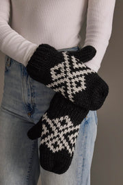 A person in a white ribbed top and blue jeans holds the Black & White Patterned Mittens from Panache Apparel Collection.