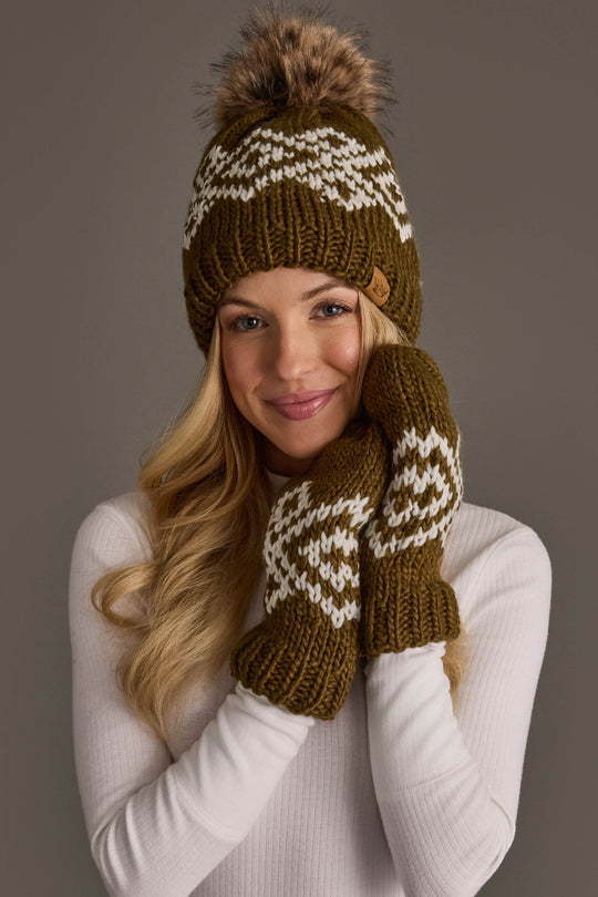 A woman smiles as she touches her cheek, wearing the Olive & White Patterned Pom Hat from Panache Apparel Collection and matching mittens. She has long blonde hair and is dressed in a white ribbed sweater.