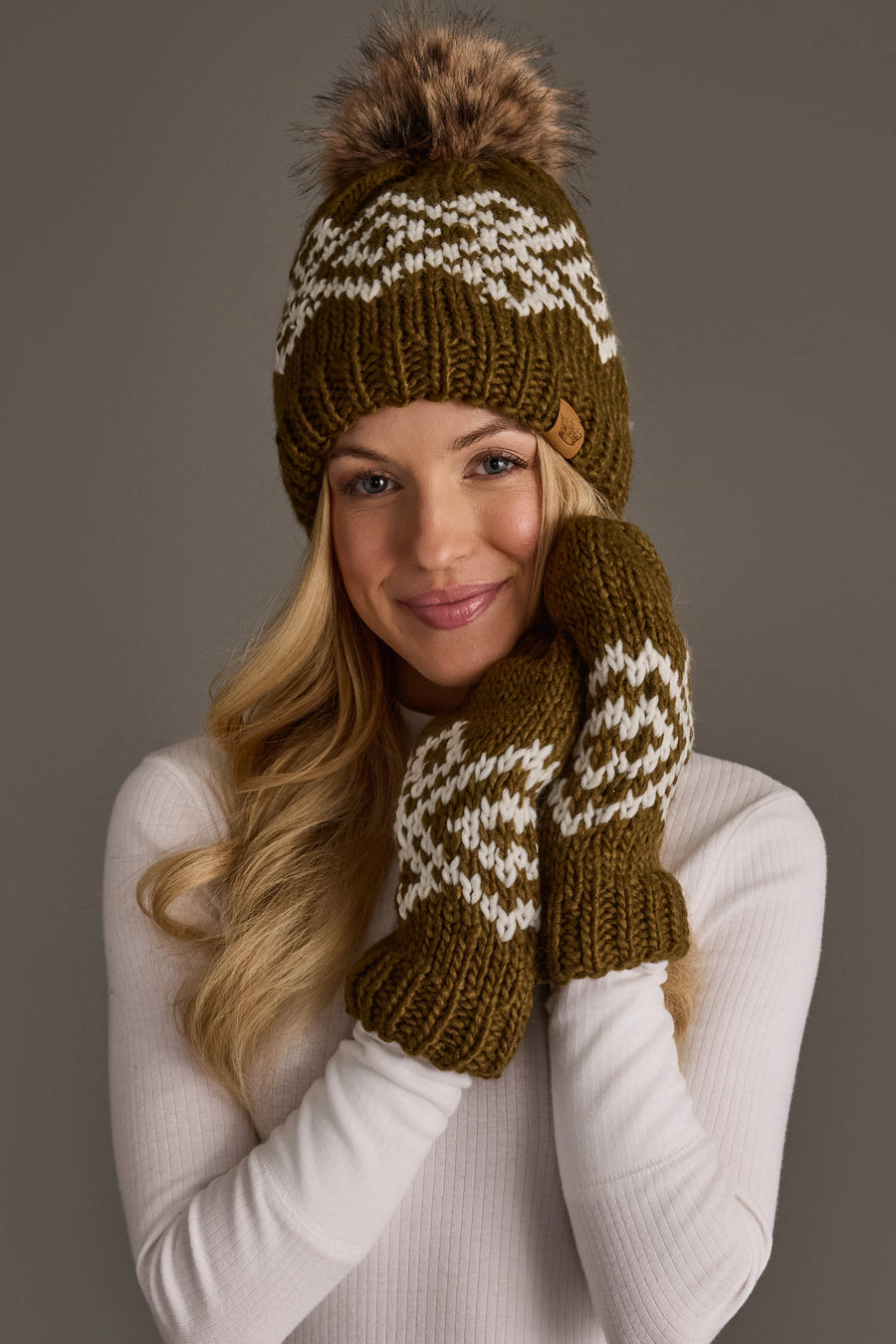 A woman smiles as she touches her cheek, wearing the Olive & White Patterned Pom Hat from Panache Apparel Collection and matching mittens. She has long blonde hair and is dressed in a white ribbed sweater.